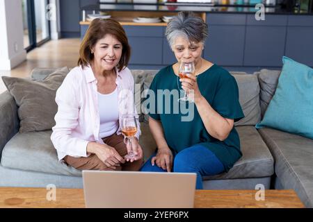 Ältere Freundinnen genießen zu Hause Wein und Videoanrufe auf dem Laptop. Freunde, Zweisamkeit, Lachen, Freizeit, Technologie, Digital Stockfoto