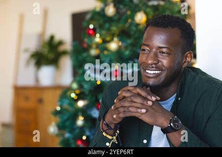 Lächelnder afroamerikanischer Mann, der Weihnachten zu Hause mit geschmücktem Baum im Hintergrund genießt. Feiern, festlich, Familie, gemütlich, Urlaub, Winter Stockfoto