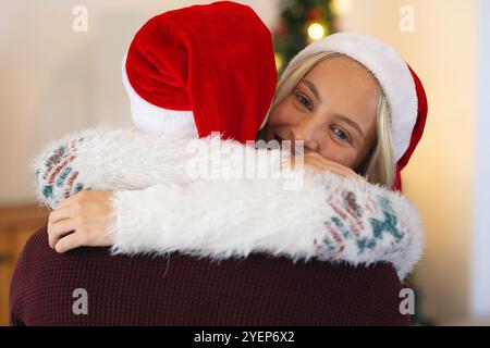 Multirassisches Paar mit Weihnachtsmützen, die sich zu Hause während der Weihnachtsfeier warm umarmen. Urlaub, fest, Liebe, Wärme, Glück, liebevoll Stockfoto
