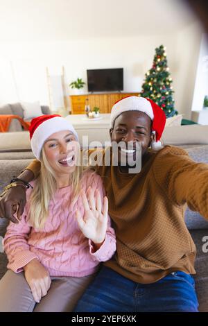 Lächelndes multirassisches Paar, das Weihnachtsmützen trägt und zu Hause Selfie macht. Feier, Feiertag, festlich, Freude, fröhlich, gemütlich Stockfoto