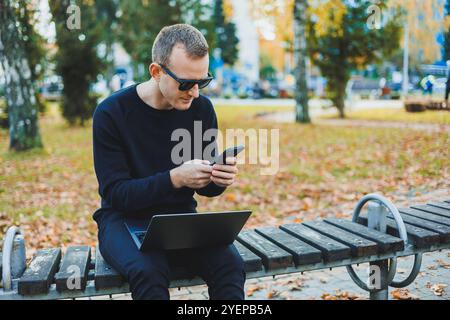 Attraktiver junger Mann in schwarzem Pullover und Sonnenbrille, sitzt in einem Herbstpark auf einer Bank und arbeitet an einem Laptop Stockfoto