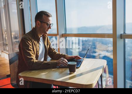 Ein ausgewogener Mann in Brille und lässiger Kleidung arbeitet an einem Laptop von einem gemütlichen Arbeitsplatz aus. Ein erfolgreicher Freelancer arbeitet Remote. Stockfoto