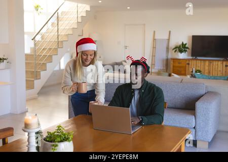 Multirassisches Paar, das zu Hause Weihnachten feiert, Laptop benutzt und Kaffee genießt. Feiertag, Feier, Familie, festlich, Winter, gemütlich Stockfoto
