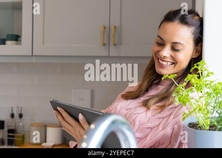 Lächelnde Frau in der Küche liest Tablette in der Nähe von Topfpflanzen, genießt Freizeit, zu Hause. Technologie, Heimarbeit, Glück, Entspannung, digital, lif Stockfoto