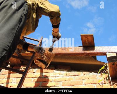 Ein Mann in Arbeitskleidung und Handschuhen malt oder bürstet eine hölzerne Dachkonstruktion gegen einen blauen Himmel, Imprägnierung von Balken und Brettern an der Basis Stockfoto