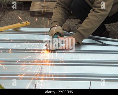 Ein Mann in Arbeitskleidung und Handschuhen schneidet oder sägt eine Metallplatte oder ein Blech, die auf dem Asphalt liegt und aus dem Funken fliegen Stockfoto