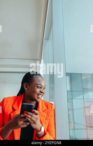 Eine fröhliche schwarze Frau in einem leuchtenden orangefarbenen Blazer steht in der Stadt an einem Bürofenster und lächelt, während sie ihr Smartphone überprüft. Ein Moment der Positivität Stockfoto