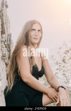 Frau, Kleid, Brille - Porträt einer Frau, die ein schwarzes Kleid und eine Brille trägt, während sie auf einem Felsen sitzt. Stockfoto