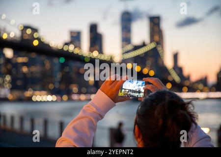 Foto einer Person, die ein Smartphone hält, um ein Foto von der Brooklyn Bridge und der Skyline von Manhattan in der Abenddämmerung zu machen. Stockfoto