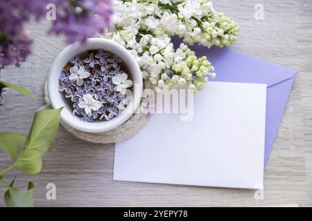 Leerer Umschlag Kopierraum Papiernote mit leckerem schwarzem Tee in weißer Tasse auf Fensterbank mit aromatischen Fliederblüten. Frühlingskomposition Tasse Fliedertee Stockfoto