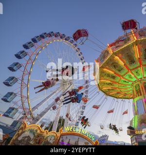 Ein Jahrmarkt in der Abenddämmerung mit beleuchtetem Kettenkarussell und Riesenrad, Europa Rad, Fahrten, Wellenflug, Cannstatter Wasen, Volksfest Bad Cannstatt, S Stockfoto