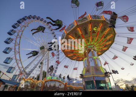 Ein Jahrmarkt in der Abenddämmerung mit beleuchtetem Kettenkarussell und Riesenrad, Europa Rad, Fahrten, Wellenflug, Cannstatter Wasen, Volksfest Bad Cannstatt, S Stockfoto