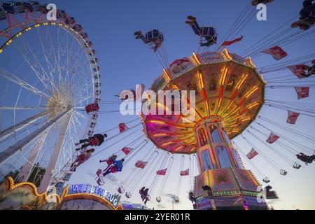 Ein Jahrmarkt in der Abenddämmerung mit beleuchtetem Kettenkarussell und Riesenrad, Europa Rad, Fahrten, Wellenflug, Cannstatter Wasen, Volksfest Bad Cannstatt, S Stockfoto