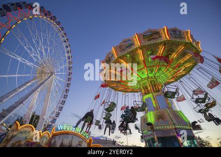 Ein Jahrmarkt in der Abenddämmerung mit beleuchtetem Kettenkarussell und Riesenrad, Europa Rad, Fahrten, Wellenflug, Cannstatter Wasen, Volksfest Bad Cannstatt, S Stockfoto