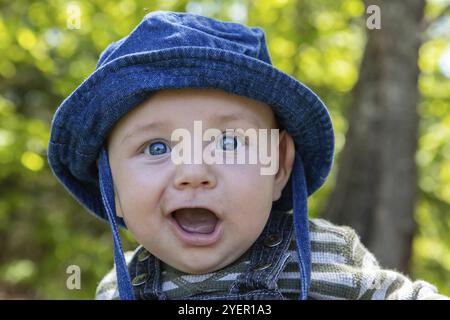 Portrait von niedlichen kleinen Säugling mit blauen Augen Hut sitzen Im Park mit weit geöffneten Mund genießen und entspannen im Freien Garten Stockfoto