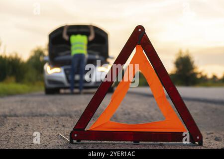 Nicht wiedererkennbarer trauriger Fahrer in reflektierender Weste. Männlicher Fahrer, der in der Nähe eines kaputten Autos steht, mit offener Motorhaube. Rotes Dreieck, um andere Verkehrsteilnehmer vor dem Auto bre zu warnen Stockfoto