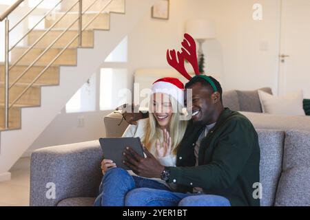 Weihnachten, multirassische Paare unterhalten sich auf Tablet, tragen festliche Hüte, lächeln zu Hause. Feiertage, Feier, Technologie, digitale Kommunikation Stockfoto