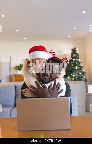 Glückliches multirassisches Paar, das zu Hause mit Laptop umgeht und gemeinsam Weihnachten feiert. Feier, Urlaub, Liebe, Zweisamkeit, Romantik, Glück Stockfoto