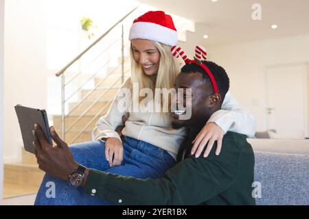 Weihnachten, glückliches multirassisches Paar Video chatten auf Tablet mit festlichen Hüten zu Hause. Feier, Technologie, virtuelle Kommunikation, freudig Stockfoto