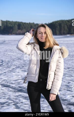 Junge Frau lächelt und schaut auf eine Kamera in einer Schneelandschaft. Happy Hipster Mädchen mit Hip Hop warme Winterkleidung. Aufenthalt auf gefrorenem Fluss, See. Stockfoto