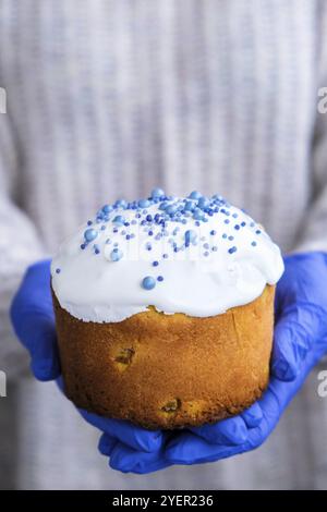 Die Hände des Kochs in blauen Handschuhen halten den Osterkuchen mit weißem Belag und blauen Streuseln. Frau mit traditionellem russischen Osterkuchen. Hausgemachter Kuchen Stockfoto