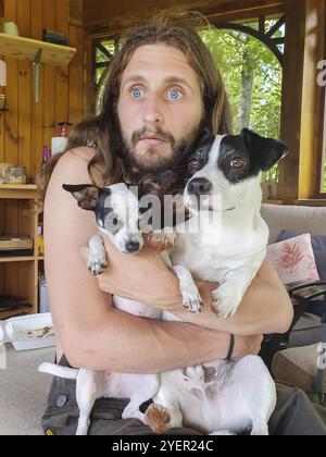 Junger Mann mit großen blauen Augen und langen blonden Haaren, die eng an der Brust halten, zwei kleine schwarze und braune Hunde, ein kleiner chihuahua und ein Terrier Stockfoto