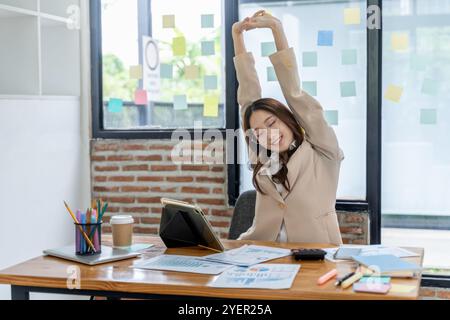 Müde junge Geschäftsfrau, die Arme und Oberkörper ausdehnt, während sie an ihrem Schreibtisch sitzt Stockfoto