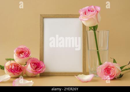 Holzrahmen mit leerem, weißem Papier und zarten Rosen auf beigefarbenem Hintergrund. Minimale trendige Komposition. Romantische pastellrosa Rosenfarbe Stockfoto