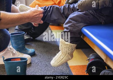 Nahaufnahme von weiblichen Händen, die dicke Wollsocken an den winzigen Füßen eines Kindergartenkindes anziehen, bevor sie ihm mit den Stiefeln helfen, nach draußen zu gehen Stockfoto