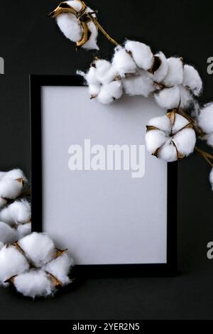 Imitierte getrocknete Baumwollblumen und einen weißen Rahmen auf einem gemütlichen schwarzen Hintergrund. Flach liegend. Speicherplatz kopieren. Bilderrahmen mit leerem Raum Stockfoto