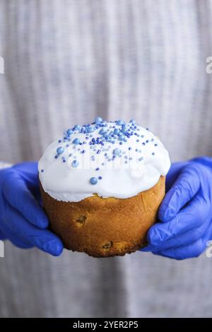 Die Hände des Kochs in blauen Handschuhen halten den Osterkuchen mit weißem Belag und blauen Streuseln. Frau mit traditionellem russischen Osterkuchen. Hausgemachter Kuchen Stockfoto