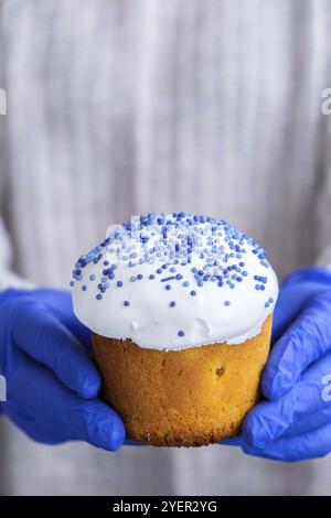 Die Hände des Kochs in blauen Handschuhen halten den Osterkuchen mit weißem Belag und blauen Streuseln. Frau mit traditionellem russischen Osterkuchen. Hausgemachter Kuchen Stockfoto