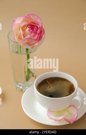 Zarte rosafarbene Rose auf Vase mit weißer Tasse Kaffee auf beigefarbenem Hintergrund. Minimale Zusammensetzung. Romantische pastellrosa Rosenblume. Moderne Ästhetik. Neutra Stockfoto