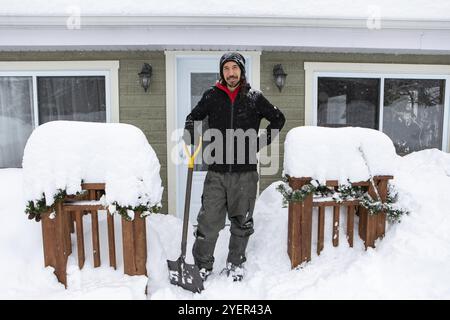 Porträt eines glücklichen kaukasischen Mannes in voller Länge, der im Freien mit einer Schaufel steht, um den Schnee zu schaufeln, der den Fußweg vor dem Haus bedeckte Stockfoto