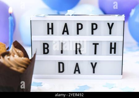 Lightbox mit Text HAPPY BIRTHDAY und Schokolade Cupcake Kerze auf Abstract unscharf festlichen Hintergrund für Urlaub. Blaue Ballons und Konfetti Stockfoto