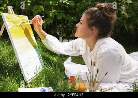 Künstler malen auf der Staffelei im Garten. Open-Air-Kunstwerkstatt. Zeichnen Sie auf der Leinwand mit Pinsel und Palette sitzen auf dem Gras Dur Stockfoto