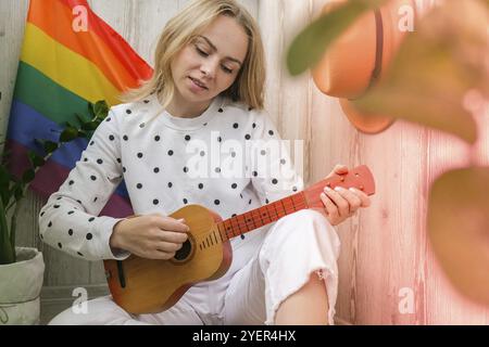 Junge Millennial-Hippie-Frau, die auf dem Balkon sitzt, spielt Gitarre. Musikunterricht und Sängersitzen. LGBTQ Regenbogenflagge auf dem Hintergrund. Online-Musikunterricht. Stockfoto