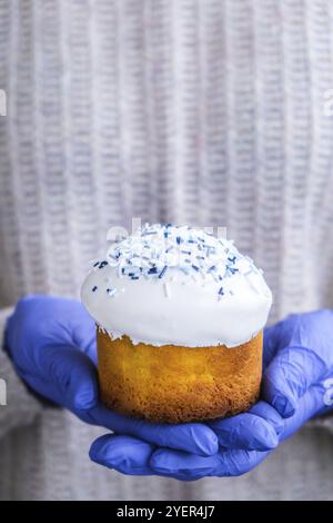 Die Hände des Kochs in blauen Handschuhen halten den Osterkuchen mit weißem Belag und blauen Streuseln. Frau mit traditionellem russischen Osterkuchen. Hausgemachter Kuchen Stockfoto