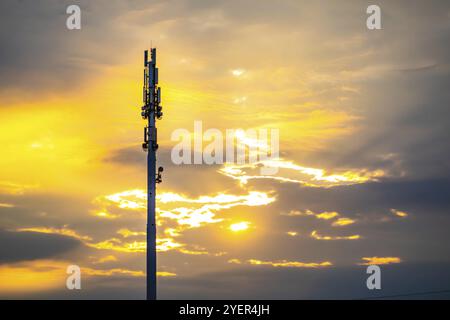 Eine Weitwinkelaufnahme eines Monopole Zelle Aufstellungsort Turm bei Sonnenuntergang. Golden beleuchtete Silhouette Mobilfunk Basisstation mit vereinzelten Wolken und Platz kopieren Stockfoto