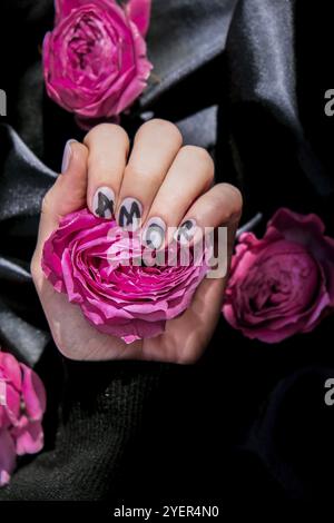 AMOR Wort auf Nägeln Maniküre halten Rosa Rose Blume auf schwarzem Seidenstoff. Liebe, Minimal flach lag Natur. Weibliche Hand. Kreative Idee. Selbstversorgung Stockfoto