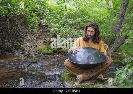 Junger Mann, der im Kreuz auf einem moosbedeckten Stein sitzt, am Ufer eines Waldflusses, der eine große Harmonie mit Gefühlen spielt Stockfoto