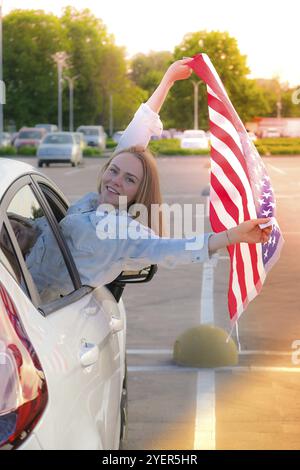 Junge, tausendjährige blonde Frau, die mit amerikanischer Flagge aus dem Auto schaut. Flagge der vereinigten Staaten in ihren Händen. 4. Juli Unabhängigkeitstag. USA Patriotismus n Stockfoto