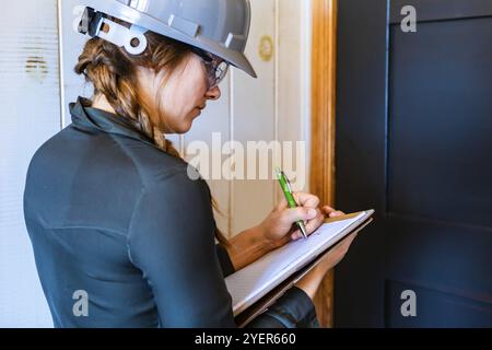 Die Arbeitnehmerin trägt einen grauen Schutzhelm und eine Schutzbrille bei der Arbeit. Eine Bauinspektorin macht sich Notizen auf ihre Zwischenablage während einer Hausinspektion Stockfoto