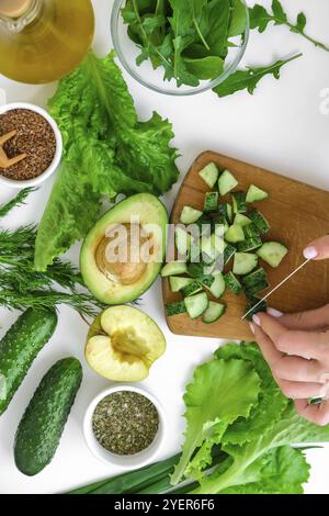 Frau kocht Salat aus frischem grünem Gemüse und Kräutern. Kochen gesunde Ernährung oder vegetarische Speisen. Weibliche Hände schneiden Gurke auf Schneidebrett surrounde Stockfoto