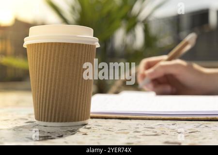 Unbekannte junge Frau studiert im Strandresort. Trinken Sie Kaffee aus einer Papiertasse. Schüler machen Hausaufgaben weibliche Hände schreiben auf Notizbuch Dankbarkeit j Stockfoto