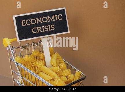 Text ZUR WIRTSCHAFTSKRISE auf dem Etikett an der Tafel mit einem Einkaufswagen gefüllt mit Pasta auf beigem Hintergrund. Einkaufs- und Lebensmittelpreise erhöhen sich Stockfoto