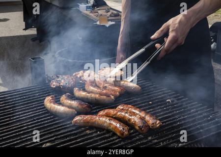 Ein Mann aus der Nähe, der mit einer Zange Würstchen über einen Grill mit steigendem Dampf dreht und warme Speisen zubereitet, die auf einer lokalen Messe serviert werden Stockfoto