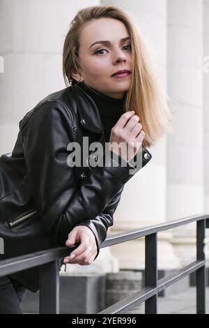 Porträt einer jungen blonden Frau mit roten Lippen in Lederjacke auf der Treppe des Firmengebäudes. Karriere Geschäftsfrau. Stylische Biker-Frau. Urban Street Stockfoto