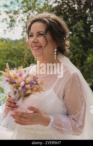 Schöne lächelnde Braut in traditionellen weißen Hochzeitskleid mit Strauß von trockenen hausgemachten Blumen. Glückliche junge Frau. Wir haben gerade geheiratet Stockfoto