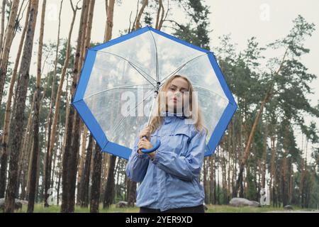 Junge Frau, die einen durchsichtigen blauen Regenschirm im Wald hält. Regnerischer Tag mit Regenschirm. Frau mit Hand, die prüft, wie lange es Rini sein wird Stockfoto